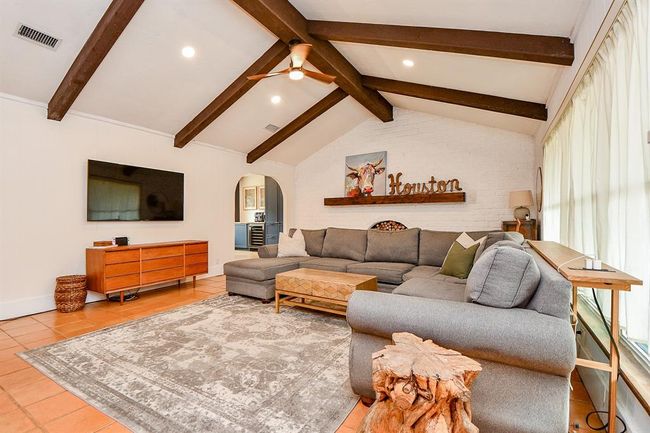 This spacious living room features a cozy sectional sofa, rustic wooden beams, and a bright atmosphere with large windows. It includes a mounted TV, stylish decor with a mix of modern and natural elements, and a decorative sign on a brick accent wall, creating a welcoming and comfortable space. | Image 11