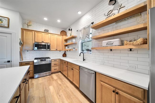 This kitchen features a modern design with light wood cabinets, open shelving, and a subway tile backsplash. It includes stainless steel appliances, ample countertop space, and a cozy window with natural light. | Image 19