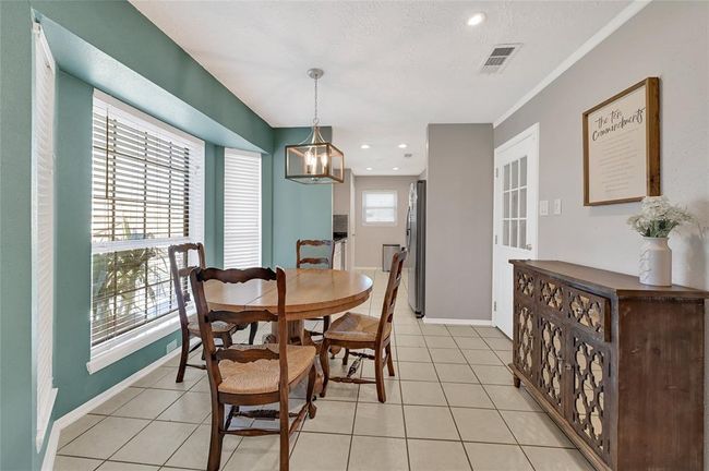 This cozy dining area features a round wooden table with chairs, large windows for natural light. The space has tile flooring and a modern light fixture, offering a welcoming atmosphere next to the kitchen. | Image 7