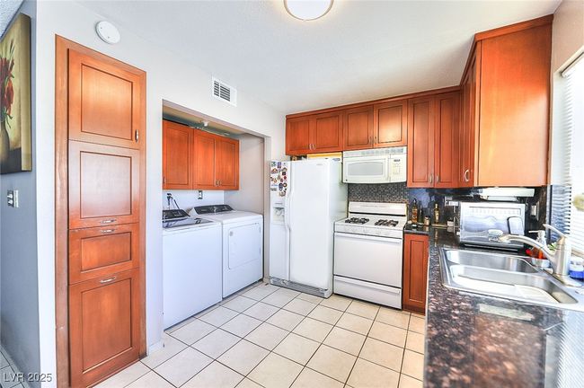 Kitchen with white appliances, washing machine and dryer, dark countertops, decorative backsplash, and light tile patterned floors | Image 5