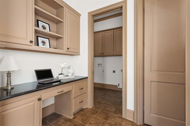 A cozy office nook with built-in cabinets and a sleek black countertop, leading into a utility room with additional cabinetry. The space has a warm, neutral color palette and tile flooring. | Image 24