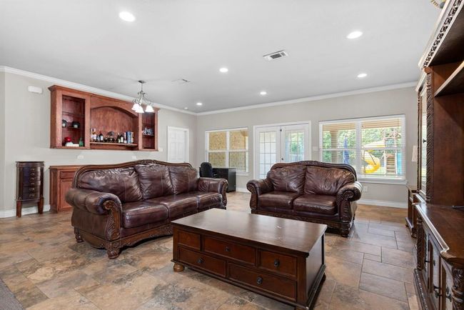 This is a spacious living room with elegant dark leather sofas, a large center table, and a built-in wooden bar area. Natural light streams in through the windows, and the room features tile flooring and recessed lighting. | Image 14