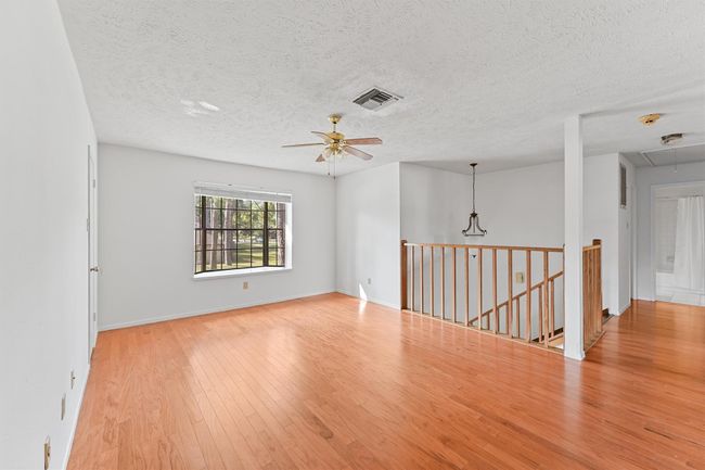 Bright upstais living space with hardwood floors, a ceiling fan, and a large window offering natural light. Features a wooden railing overlooking a staircase, creating an open, airy feel. Perfect for a cozy and inviting home. | Image 16
