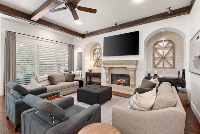 The Living room features a cedar beam ceiling and crown molding, wood shutters, solid wood floors, ceiling fan, arched walkways, and a cast stone gas log fireplace | Image 8