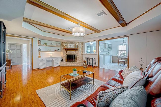 This spacious living room features a cozy brick fireplace, rich wood flooring, and elegant coffered ceilings with wood accents. It seamlessly connects to the kitchen and dining area through an open layout, creating a welcoming space for entertaining. | Image 8