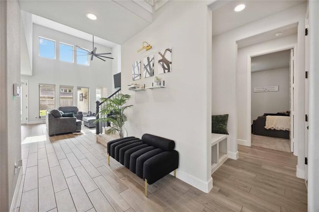 This home features a bright, open living area with high ceilings and ample natural light from large windows. The modern design includes wood-look tile flooring, neutral walls, and sleek furnishings. A cozy hallway leads to additional rooms, adding to the spacious feel. | Image 8