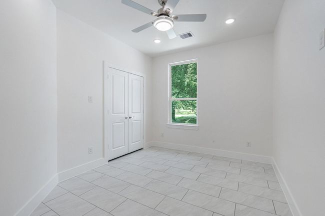 This room features a clean, modern design with white walls and tiled flooring. It includes a ceiling fan, recessed lighting, a window providing natural light, and a closet with double doors. Perfect for a bedroom or home office. | Image 21
