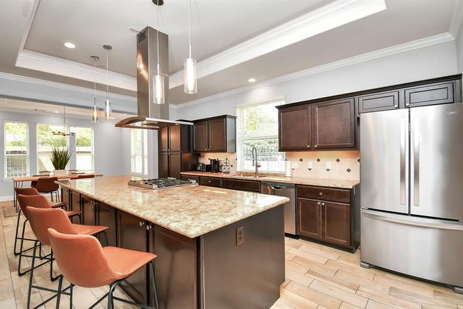 This modern kitchen features a spacious island with granite countertops, stylish pendant lighting, and sleek stainless steel appliances. The dark wood cabinetry complements the neutral tones, and there's ample natural light from large windows, creating a welcoming and functional space. | Image 9