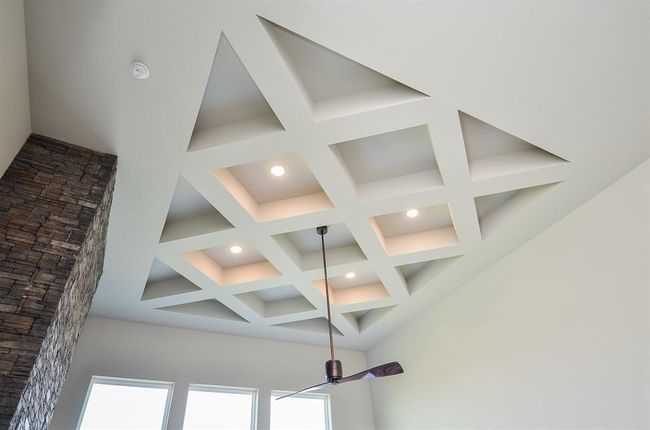 This photo showcases a modern ceiling with a coffered design, featuring recessed lighting and a sleek ceiling fan. A stone accent wall adds texture and elegance, while large windows provide natural light. | Image 9