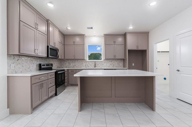 This modern kitchen features taupe cabinetry, a spacious island, and sleek stainless steel appliances. The light tile flooring and herringbone backsplash add a stylish touch, while recessed lighting keeps the space bright and inviting. | Image 16