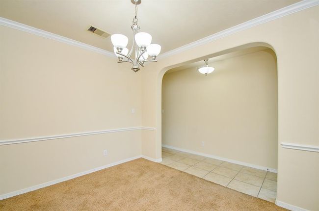 This photo shows a bright dining area with soft beige walls, a plush carpet, and a stylish chandelier. An adjacent space with tiled flooring features a matching light fixture, separated by an arched entryway, enhancing the elegant open layout. | Image 8