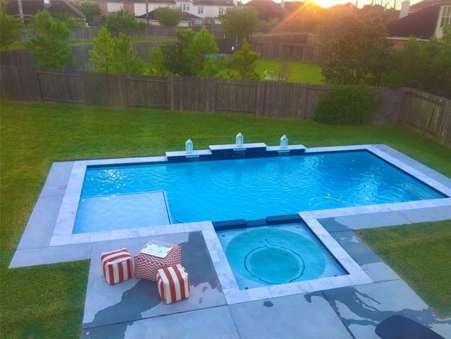 This backyard features a modern rectangular pool with a connected circular hot tub, surrounded by a spacious patio. The area includes a grassy lawn and is enclosed by a wooden fence, offering privacy and a serene atmosphere. | Image 5