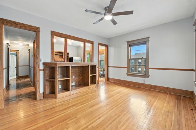 Sun-filled living area with gleaming hardwood floors, ceiling fan, and built-in wood shelving open to the kitchen. | Image 9