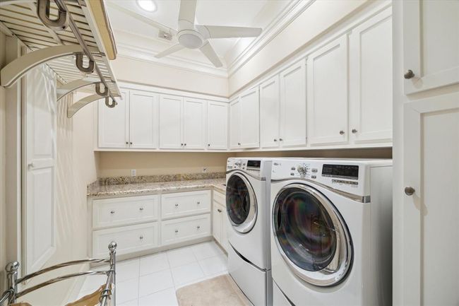 This laundry room features plenty of overhead cabinets for storage, granite countertops, and a ceiling fan for ventilation. Bright and functional with white cabinetry and tile flooring. | Image 29