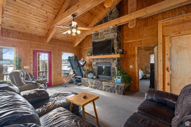 Carpeted living room featuring beam ceiling, ceiling fan, a stone fireplace, wooden walls, and wood ceiling | Image 22