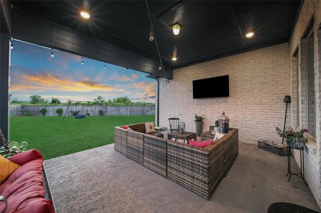 This photo showcases a spacious covered patio with comfortable seating and a mounted TV, perfect for outdoor relaxation. The area opens to a large, well-maintained backyard with a swing set, all enclosed by a wooden fence, providing privacy and a great space for family activities. | Image 6