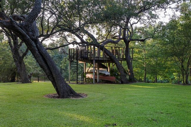 Perched amidst three majestic oak trees, a charming 16'x16' treehouse and encircling deck command picturesque views of the yard and the serene Carancahua River. Floodlights thoughtfully positioned within the treehouse cast a gentle radiance toward the river, enhancing the nighttime ambiance. Designed with both leisure and functionality in mind, racks have been ingeniously integrated underneath to securely stow canoes and rowboats. | Image 8