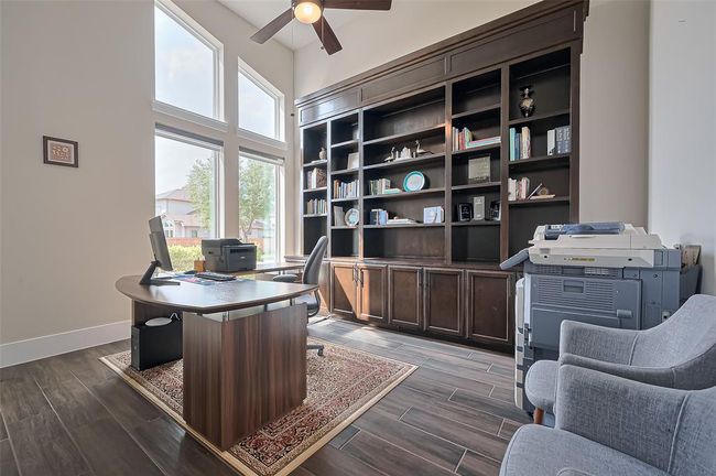This photo showcases a bright home office with large windows, built-in bookshelves, and a stylish wooden desk. The high ceiling and fan add to the spacious feel, while the dark wood floors and area rug provide warmth. | Image 6