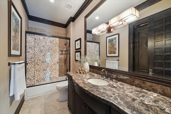 One of five guest bathrooms, this refined bathroom pairs warm tones with polished finishes, creating a stylish and welcoming atmosphere. A richly veined granite countertop adds a sense of luxury, while framed mirrors and contemporary lighting enhance the space’s elegance. The tub/shower combination features detailed tilework and a bold patterned glass enclosure, offering both function and flair. Ideal for guests, it delivers comfort with an upscale touch. | Image 32