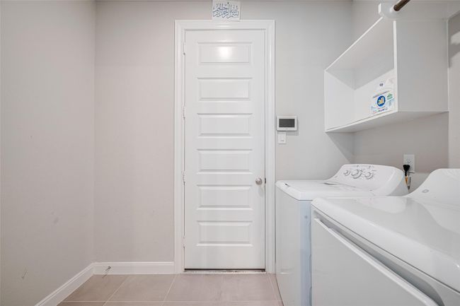 This photo shows a clean, modern laundry room with a washer and dryer set, white shelving for storage. The space is functional with tile flooring and a door leading to the garage. | Image 22