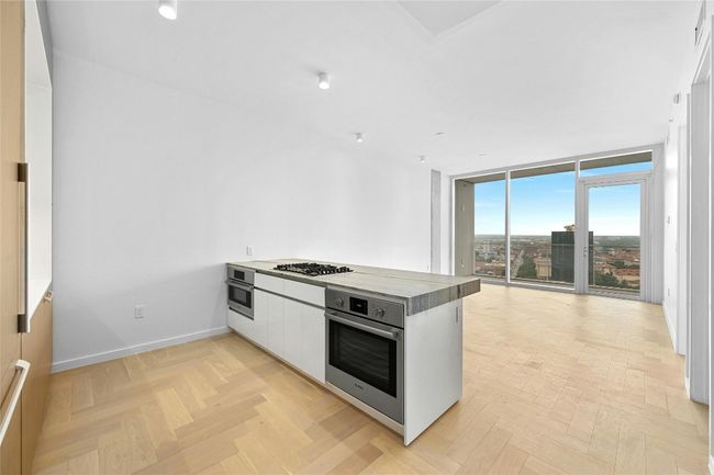 A modern, open-concept kitchen, dining and living area. The kitchen features a sleek, minimalist design with a large island at its center, topped with a light-colored stone countertop. The island includes a built-in gas cooktop and stainless steel oven. | Image 4