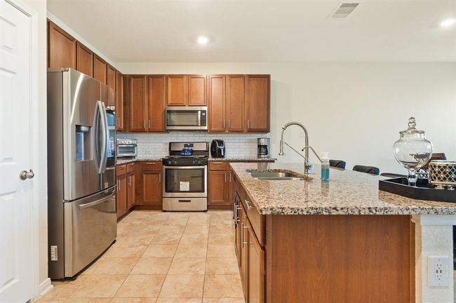 This kitchen features modern stainless steel appliances, warm wooden cabinetry, and a spacious granite-topped island with a built-in sink, perfect for cooking and entertaining. The tiled floor and neutral backsplash add a clean, contemporary touch. | Image 14