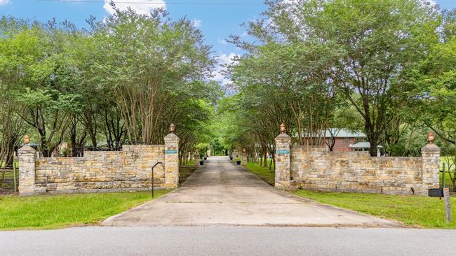 A welcoming entrance features a wide driveway flanked by stone walls and lush trees, leading to a secluded home in a serene setting. | Image 6