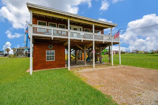 This elevated two-story home features a spacious balcony, a wooden exterior, and a covered area underneath perfect for parking or outdoor activities. Surrounded by open green space, it offers a serene setting with a coastal vibe. | Image 31