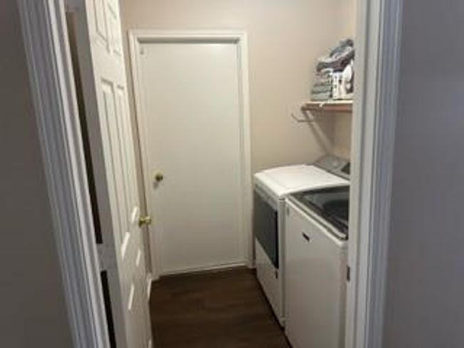This photo shows a compact laundry room with a washer and dryer side by side. There's a shelf above for storage, and the room features a neutral color scheme with wooden flooring. | Image 4