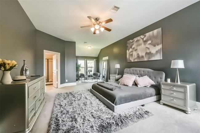 Spacious, elegant bedroom with plush carpeting and soft gray walls. Features ceiling fan and ample natural light from a windowed seating area. | Image 18