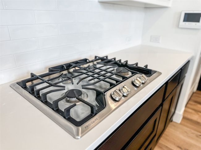 Kitchen view of stainless steel gas stovetop, backsplash, light countertops, light wood-type flooring, and dark brown cabinetry | Image 14