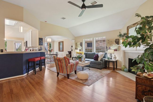 This inviting living space features an open layout with warm wood flooring, a cozy sitting area with a fireplace, and abundant natural light from large windows. The kitchen and dining area are seamlessly integrated, offering a welcoming atmosphere for entertaining. | Image 5