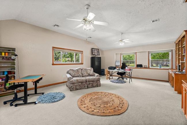 This spacious living area features built-in wooden shelving, a cozy seating arrangement, and ample natural light from large windows overlooking a lush green yard. Equipped with ceiling fans, it combines functionality with a peaceful view. | Image 13