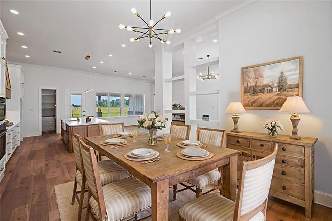 This spacious, open-concept kitchen and dining area features elegant lighting, a rustic wooden dining table, and a large island with a sink. The room is bright with natural light from large windows, offering a view of the outdoors. The decor is warm and inviting, with LVP floors and tasteful artwork. DIGITALLY STAGED | Image 11