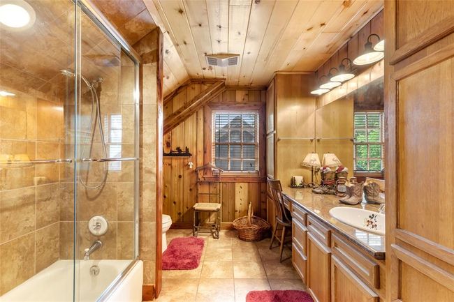 This rustic bathroom features warm wood paneling, a glass-enclosed shower-tub combo, and ample storage with built-in cabinets. It has a cozy, cabin-like atmosphere with natural light from a window above the sink area. | Image 43