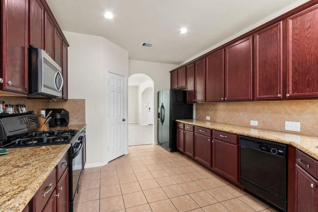This kitchen features dark wood cabinetry, granite countertops, and stainless steel appliances. It has a spacious layout with tiled flooring and an arched entryway leading to another room. | Image 10
