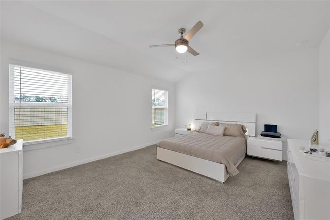 This spacious bedroom features a neutral color palette with carpeted flooring, a ceiling fan, and ample natural light from two windows. It includes a comfortable bed with matching nightstands, creating a modern and inviting atmosphere. | Image 12
