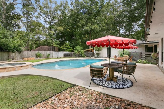 This backyard features a spacious pool with a built-in hot tub, surrounded by a patio with seating and a red umbrella, all set against a lush, wooded backdrop for privacy. | Image 43
