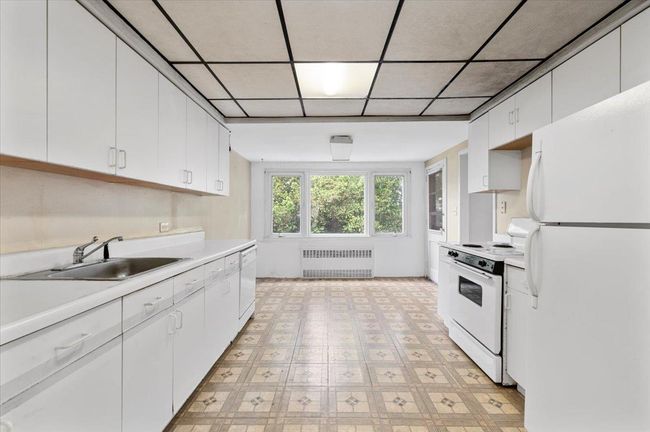 Kitchen with white appliances, radiator heating unit, light countertops, a sink, and white cabinetry | Image 6