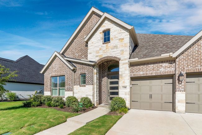 French country inspired facade featuring a front lawn, brick siding, concrete driveway, stone siding, and a shingled roof | Image 4