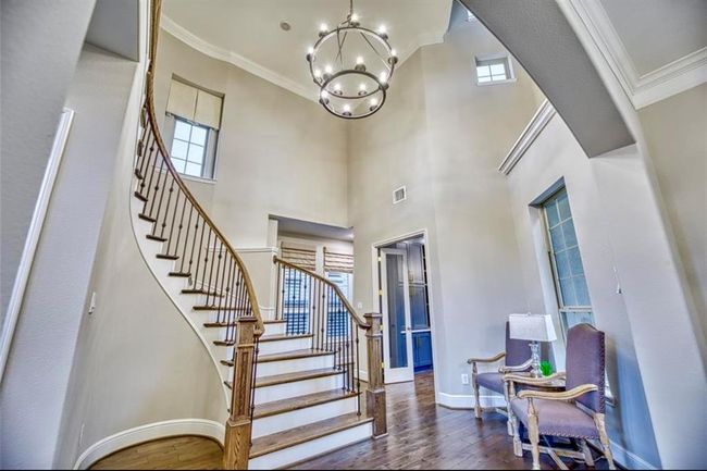 This photo showcases an elegant entryway featuring a grand, curved staircase with wooden steps and wrought iron railing. The space is bright and airy, with high ceilings and a stylish chandelier. Adjacent to the staircase, there is a cozy sitting area with two armchairs and a lamp, adding a welcoming touch. | Image 10