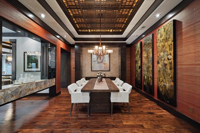 This designer dining room exudes modern sophistication with layered textures, warm wood tones, and museum-quality finishes. A geometric multi-layered coffered ceiling in richly stained wood adds visual depth, which is beautifully illuminated by a contemporary modern chandelier. Imported European wall coverings, bold oversized art panels, and a mother of pearl accent wall create a space that is perfect for formal entertaining or elevated everyday dining. | Image 9
