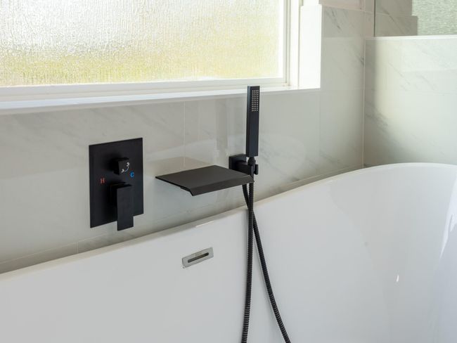 Relax and unwind in this deep soaking tub complete with a matte black wall-mounted spout and handheld shower—perfectly placed beneath a privacy window for natural light and tranquility. | Image 25