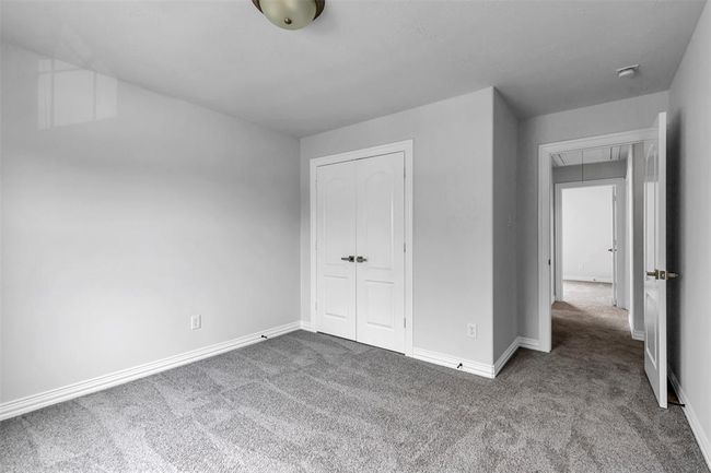 This photo shows a bright, carpeted bedroom with neutral gray walls. It features a ceiling light fixture and has a double door closet. There is an adjoining doorway leading to another room. | Image 33