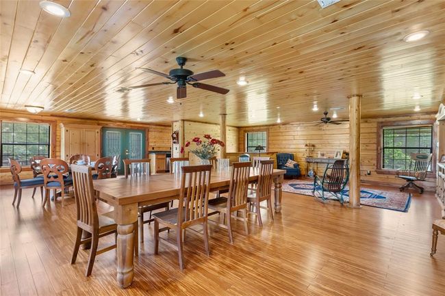 This spacious, rustic interior features wooden walls and ceiling, creating a cozy, cabin-like atmosphere. It includes a large dining area with ample natural light, a sitting area with a rug, and elegant wooden furniture, perfect for family gatherings and entertaining. | Image 49