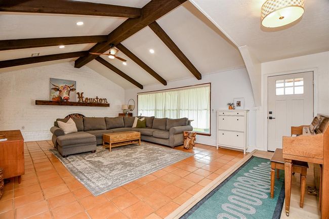 This inviting living room features a high ceiling with exposed wooden beams, a cozy sectional sofa, and warm terracotta tile flooring. A large window allows natural light, complementing the white brick accent wall and creating a bright, open space for relaxation. | Image 10