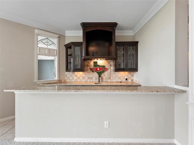 Kitchenette featuring dark brown cabinets, decorative backsplash, glass insert cabinets, crown molding, and light stone countertops | Image 32