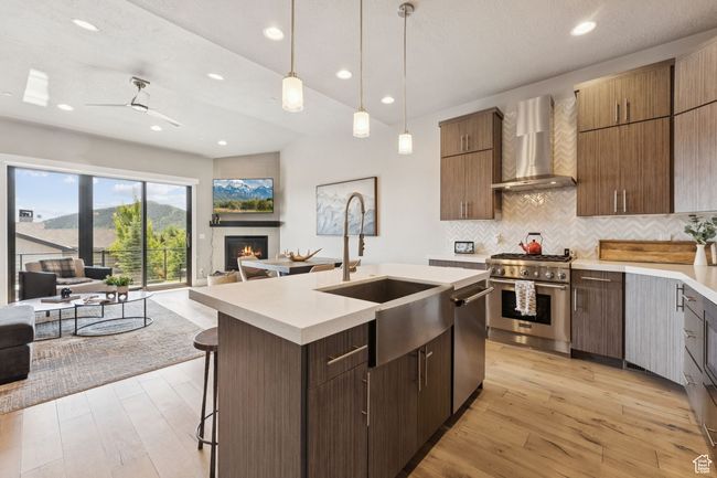Kitchen featuring wall chimney range hood, stainless steel appliances, open floor plan, light countertops, and a glass covered fireplace | Image 8