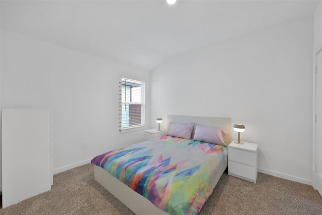 This is a bright, modern bedroom with white walls, a cozy carpet, and a colorful geometric-patterned bedspread. It features a bed with two bedside tables, each with a lamp, and a window providing natural light. | Image 17