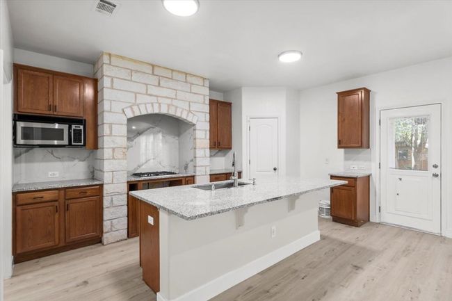 Kitchen with tasteful backsplash, light wood-style flooring, a center island with sink, light stone countertops, and brown cabinetry | Image 9